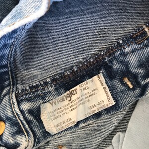 May include: A close-up of a blue denim Wrangler jeans label with white stitching. The label has the text "Wrangler 921 LBVW 7 REG." and instructions for washing and care. The label also includes the text "Made in USA 100% Cotton" and the numbers "A335-025 0 84084 39435 40".