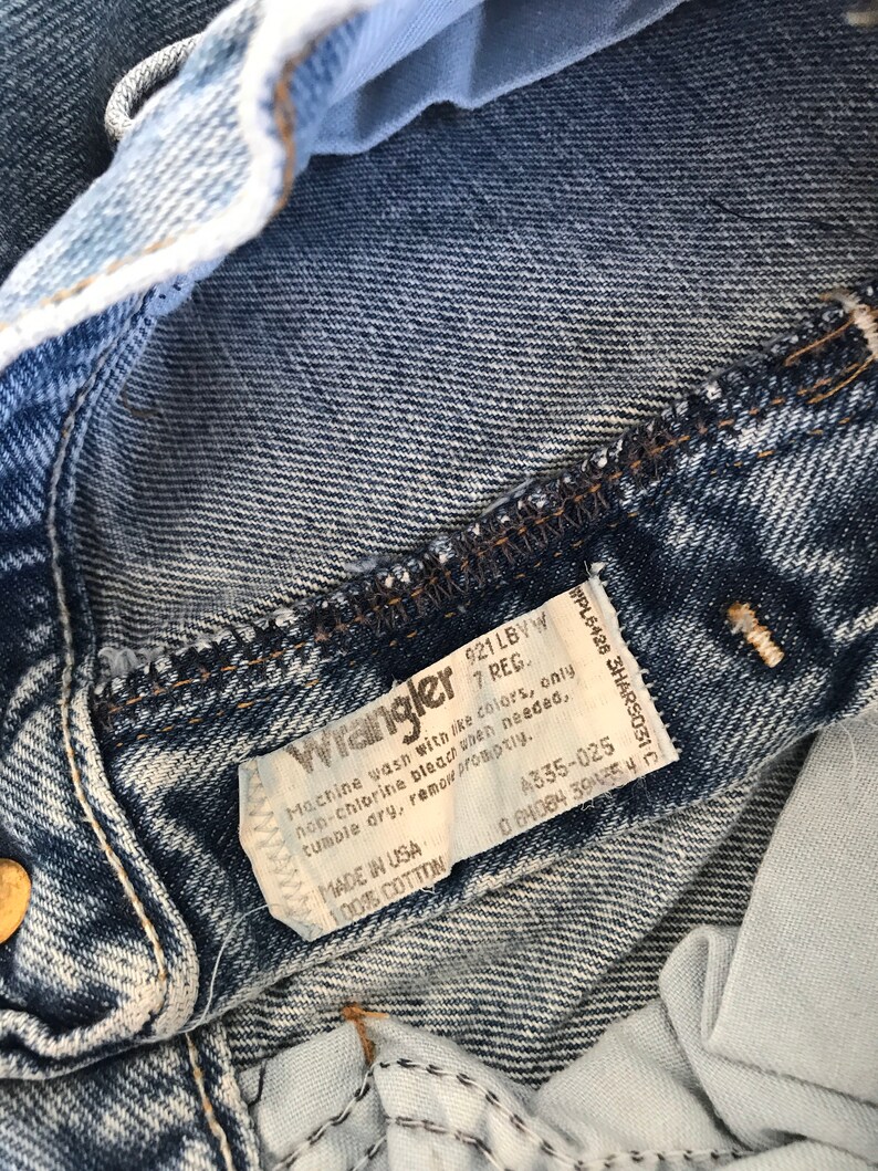 May include: A close-up of a blue denim Wrangler jeans label with white stitching. The label has the text "Wrangler 921 LBVW 7 REG." and instructions for washing and care. The label also includes the text "Made in USA 100% Cotton" and the numbers "A335-025 0 84084 39435 40".