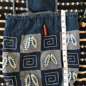 Puede incluir: Una bolsa de cord&oacute;n de mezclilla azul con una solapa superior y un cierre de bot&oacute;n de madera. La bolsa est&aacute; decorada con un dise&ntilde;o de patchwork de cuadrados azules y blancos con hojas bordadas y formas geom&eacute;tricas.