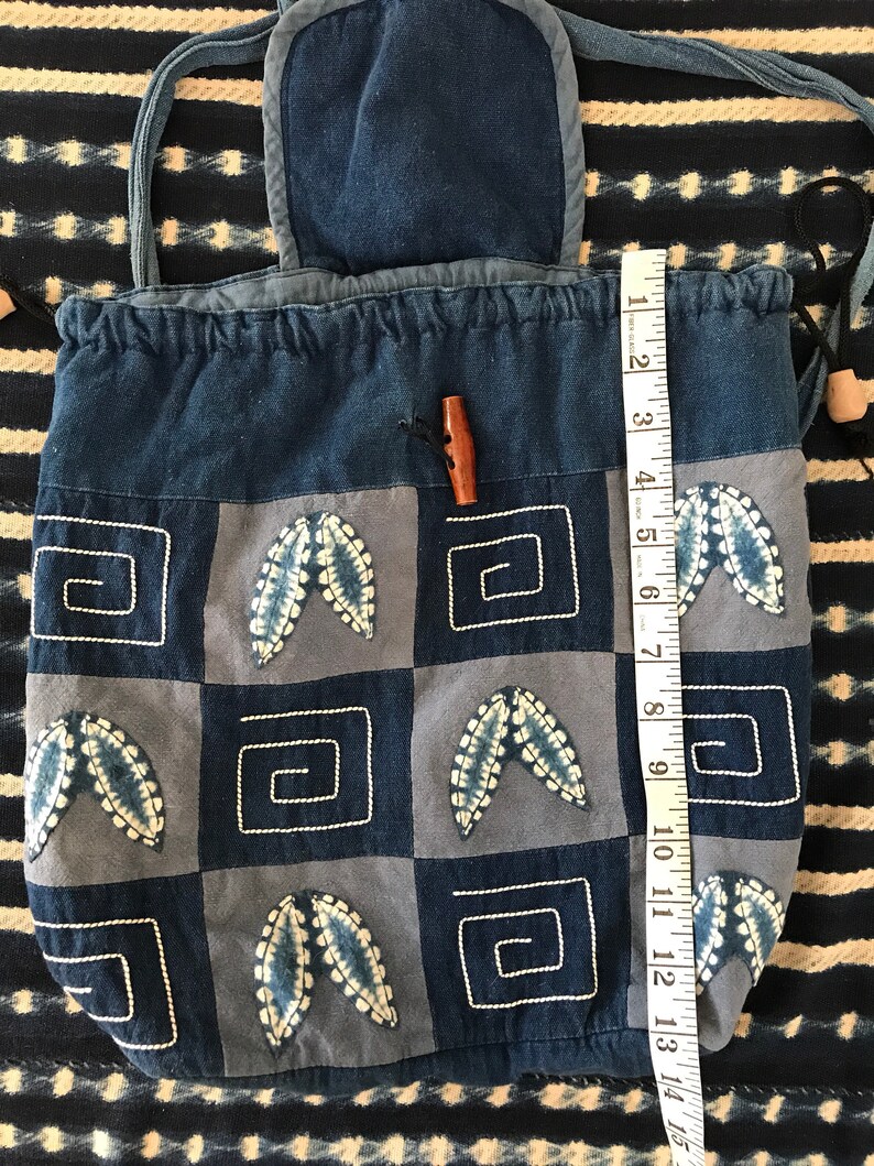 Puede incluir: Una bolsa de cord&oacute;n de mezclilla azul con una solapa superior y un cierre de bot&oacute;n de madera. La bolsa est&aacute; decorada con un dise&ntilde;o de patchwork de cuadrados azules y blancos con hojas bordadas y formas geom&eacute;tricas.