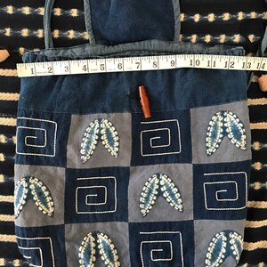 Puede incluir: Una bolsa de algod&oacute;n azul con cord&oacute;n de ajuste, tapa con solapa y cierre de bot&oacute;n de madera. La bolsa est&aacute; decorada con un dise&ntilde;o de patchwork de cuadrados azules y blancos con hojas bordadas y formas geom&eacute;tricas.