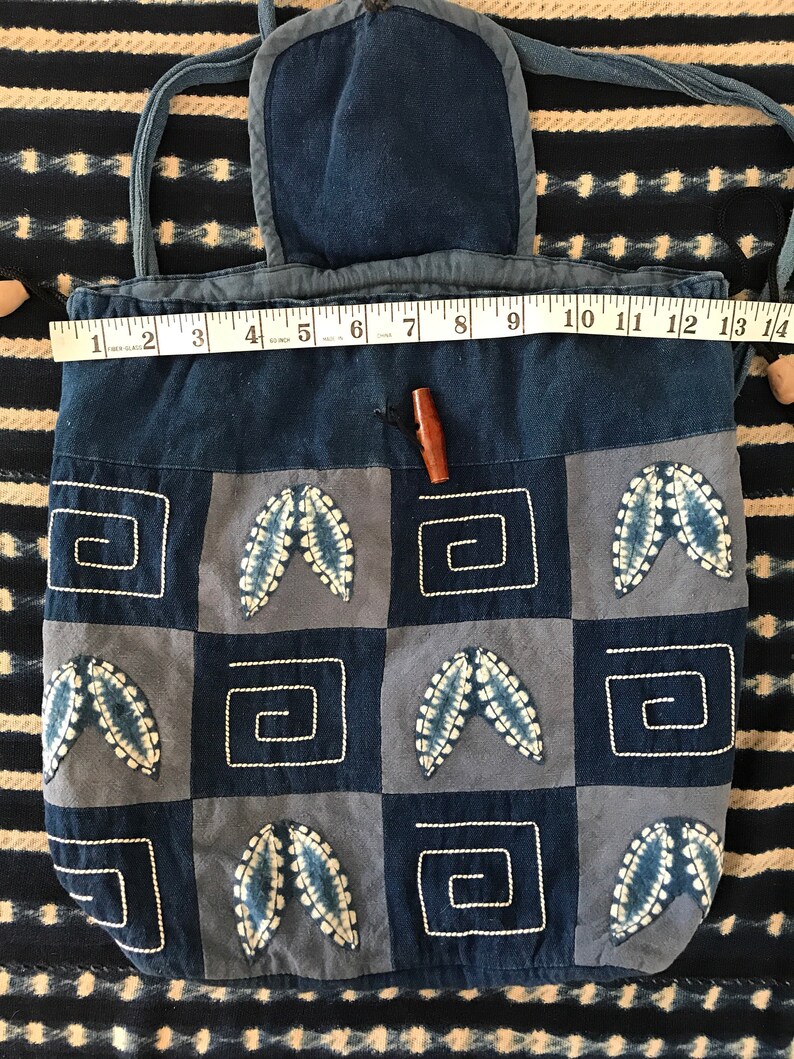 Puede incluir: Una bolsa de algod&oacute;n azul con cord&oacute;n de ajuste, tapa con solapa y cierre de bot&oacute;n de madera. La bolsa est&aacute; decorada con un dise&ntilde;o de patchwork de cuadrados azules y blancos con hojas bordadas y formas geom&eacute;tricas.