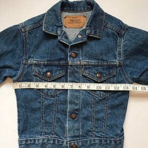 K&ouml;nnte beinhalten: Eine blaue Jeansjacke mit einem braunen Lederetikett, auf dem "Levi Strauss & Co." steht. Die Jacke hat einen Knopfverschluss und f&uuml;nf Kn&ouml;pfe auf der Vorderseite. Die Jacke wird mit einem Ma&szlig;band &uuml;ber die Brust gemessen, wobei eine Messung von 38 cm angezeigt wird.