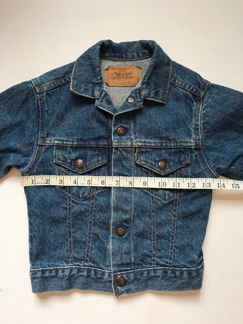 K&ouml;nnte beinhalten: Eine blaue Jeansjacke mit einem braunen Lederetikett, auf dem "Levi Strauss & Co." steht. Die Jacke hat einen Knopfverschluss und f&uuml;nf Kn&ouml;pfe auf der Vorderseite. Die Jacke wird mit einem Ma&szlig;band &uuml;ber die Brust gemessen, wobei eine Messung von 38 cm angezeigt wird.