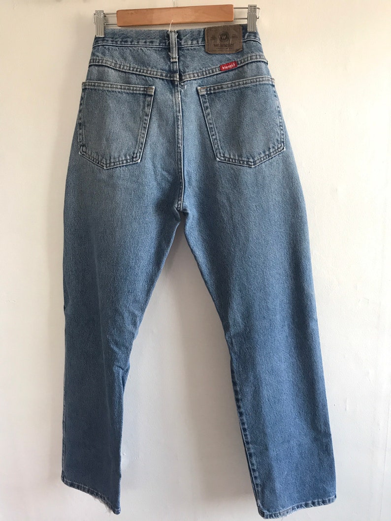 Pu&ograve; includere: Un paio di jeans in denim blu chiaro con un'etichetta Wrangler in pelle marrone sulla tasca posteriore.