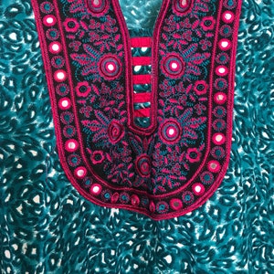 Puede incluir: Una t&uacute;nica de color azul turquesa y blanco con un cuello bordado en rosa y negro con peque&ntilde;os espejos redondos.