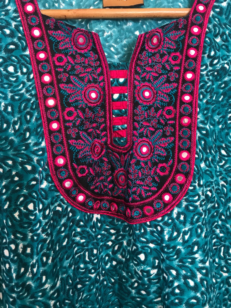 Puede incluir: Una t&uacute;nica de color azul turquesa y blanco con un cuello bordado en rosa y negro con peque&ntilde;os espejos redondos.