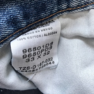 Pu&ograve; includere: Un'etichetta bianca con testo nero, che mostra la taglia e il numero di stile di un paio di jeans. L'etichetta riporta "96501DS 9650PDS 33 X 32 TZS-D187-032 244084708048".