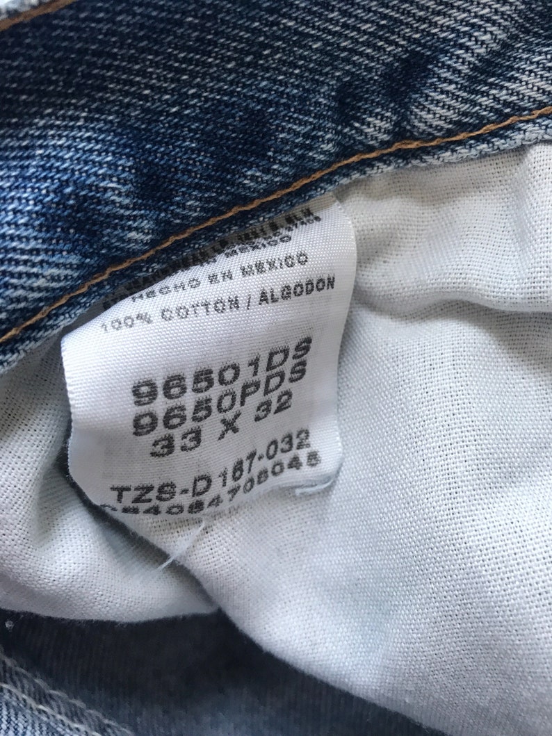 Pu&ograve; includere: Un'etichetta bianca con testo nero, che mostra la taglia e il numero di stile di un paio di jeans. L'etichetta riporta "96501DS 9650PDS 33 X 32 TZS-D187-032 244084708048".