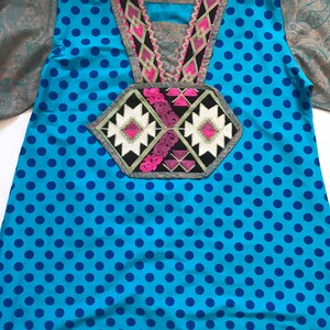 以下が含まれることがあります： 青と白のドット柄のチュニック。カラフルな刺繍のネックラインと、前面に幾何学模様が施されています。