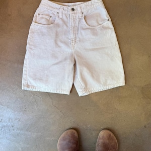 May include: Light beige denim shorts with a button closure and pockets. The shorts are high-waisted and have a classic, casual style. The image also shows brown leather clogs.
