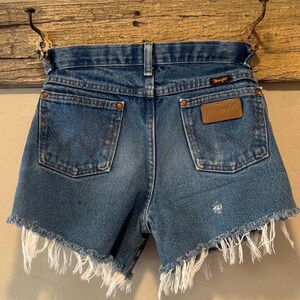 May include: Blue denim shorts with a frayed, raw-edge hem. The shorts feature two back pockets with copper-colored rivets, a brown leather Wrangler brand patch on the right pocket, and a small black Wrangler label on the waistband.