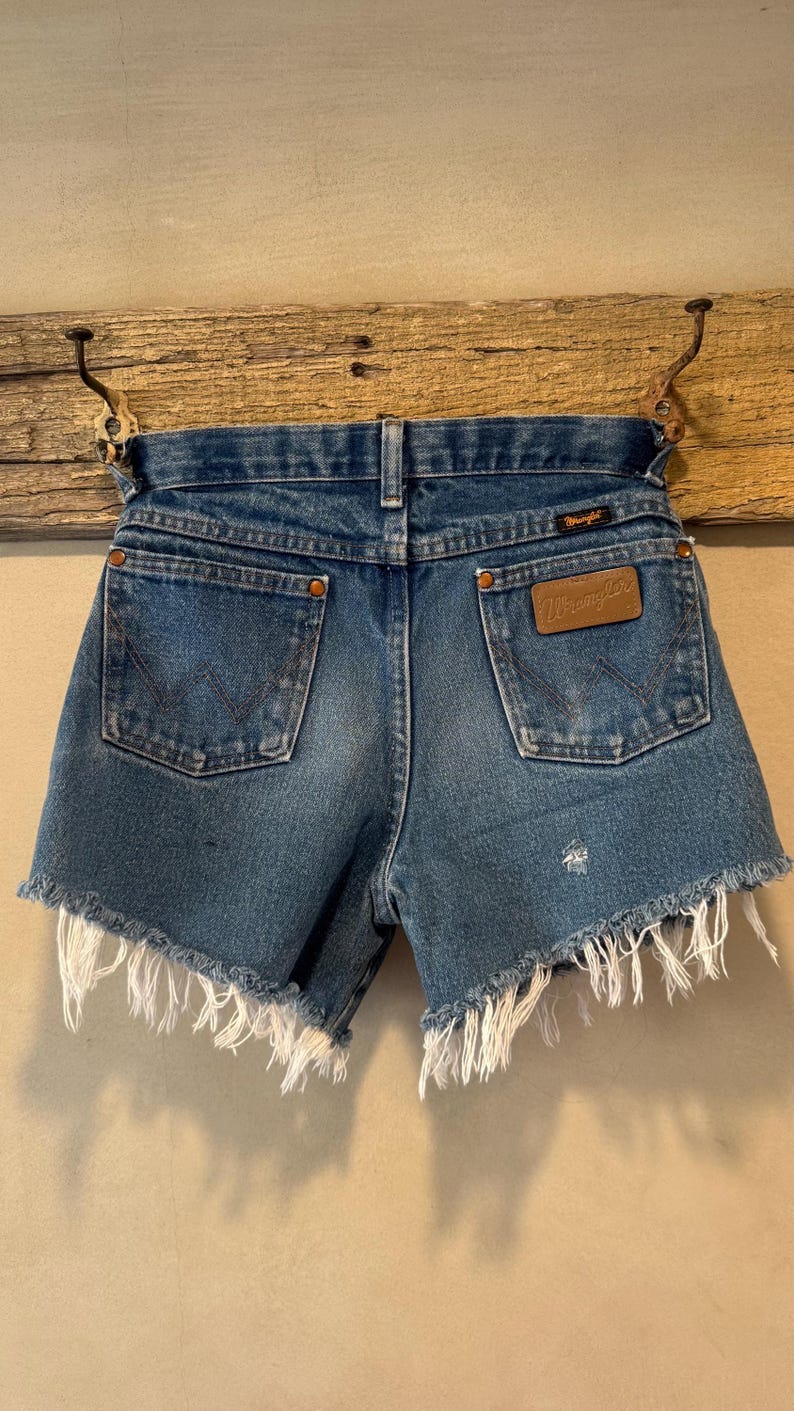 May include: Blue denim shorts with a frayed, raw-edge hem. The shorts feature two back pockets with copper-colored rivets, a brown leather Wrangler brand patch on the right pocket, and a small black Wrangler label on the waistband.