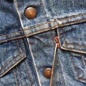 K&ouml;nnte beinhalten: Nahaufnahme einer blauen Jeansjacke mit einem kupfernen Knopf und einem braunen Lederanh&auml;nger mit der Aufschrift "Levi's" und einer Schere.