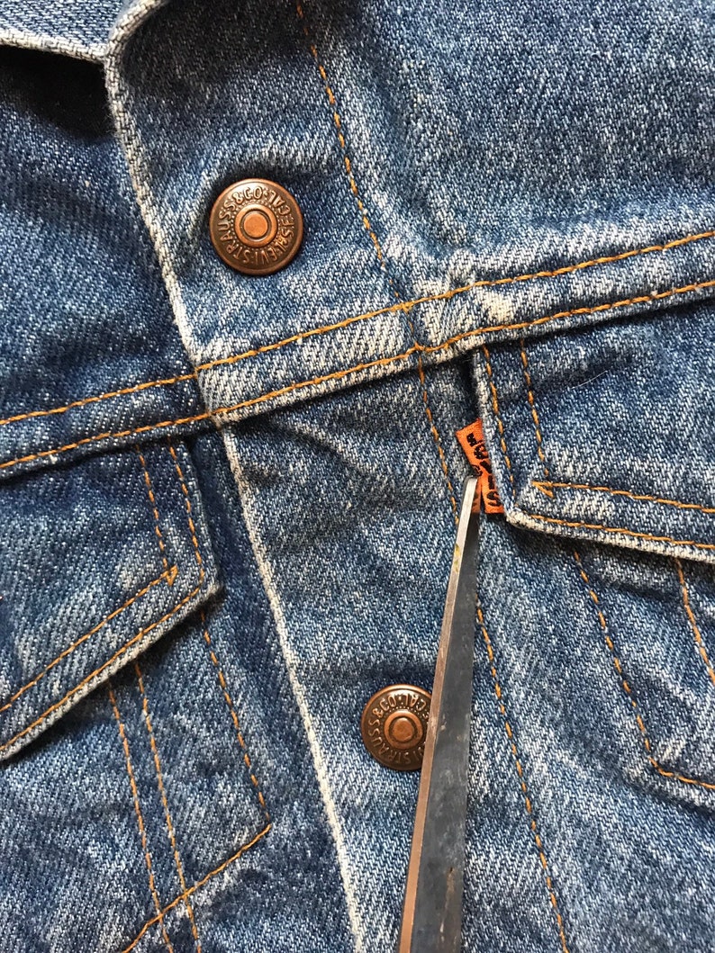 K&ouml;nnte beinhalten: Nahaufnahme einer blauen Jeansjacke mit einem kupfernen Knopf und einem braunen Lederanh&auml;nger mit der Aufschrift "Levi's" und einer Schere.
