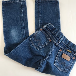 May include: Blue denim jeans with the Wrangler brand label on the back pocket. The jeans have a classic five-pocket design, copper rivets, and a straight leg. The jeans are folded on a white surface.