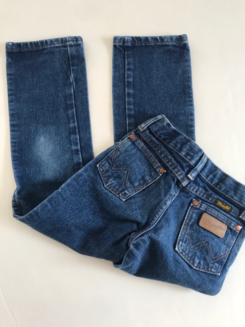 May include: Blue denim jeans with the Wrangler brand label on the back pocket. The jeans have a classic five-pocket design, copper rivets, and a straight leg. The jeans are folded on a white surface.