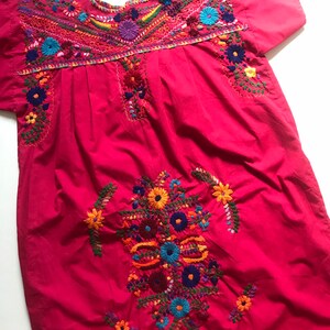 May include: A pink cotton dress with colourful floral embroidery. The dress has a round neckline and short sleeves.