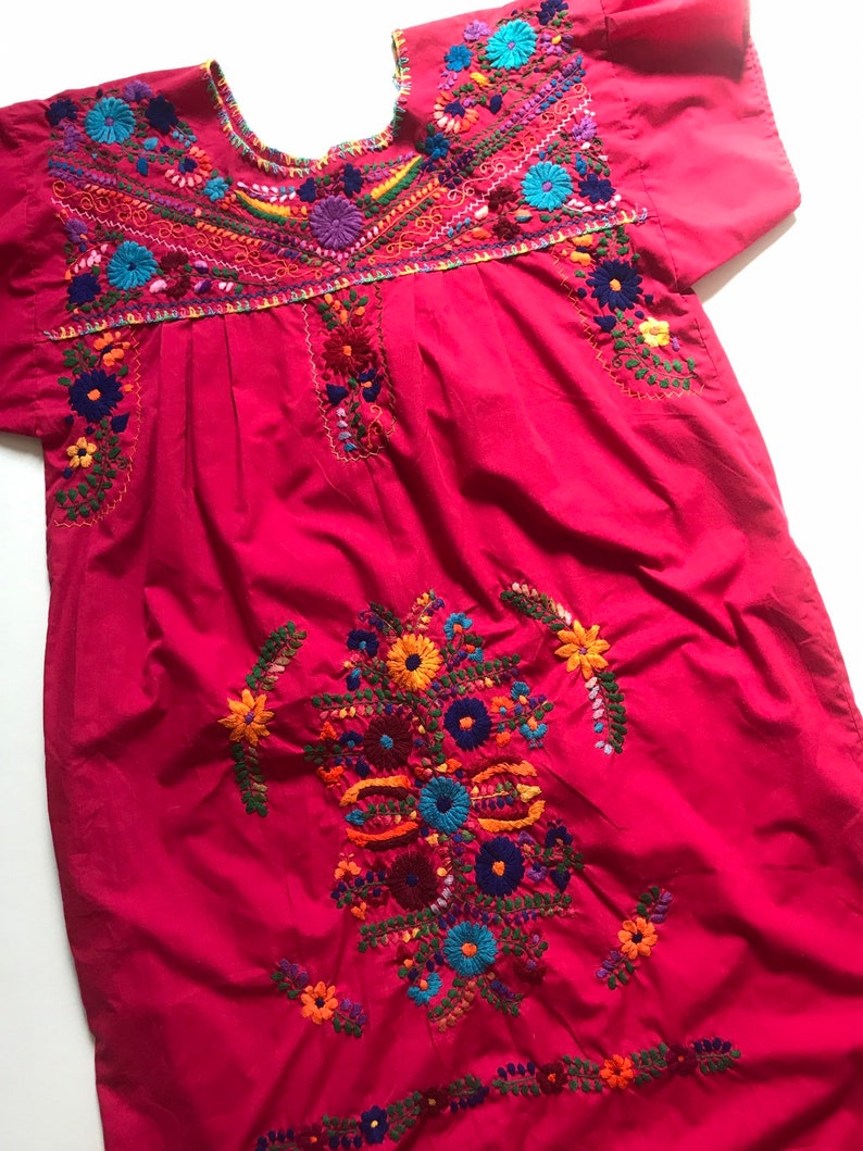 May include: A pink cotton dress with colourful floral embroidery. The dress has a round neckline and short sleeves.