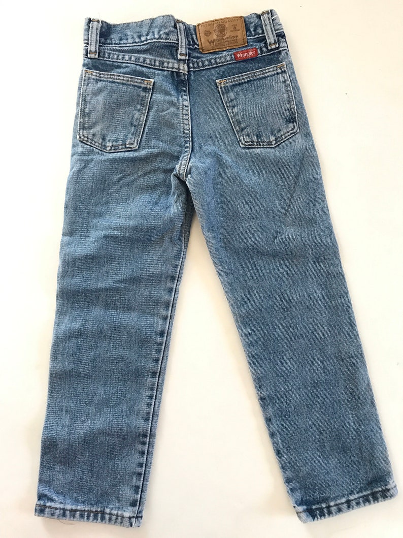 May include: A pair of light blue denim jeans with a brown leather patch on the back pocket that says "Wrangler" in white text.