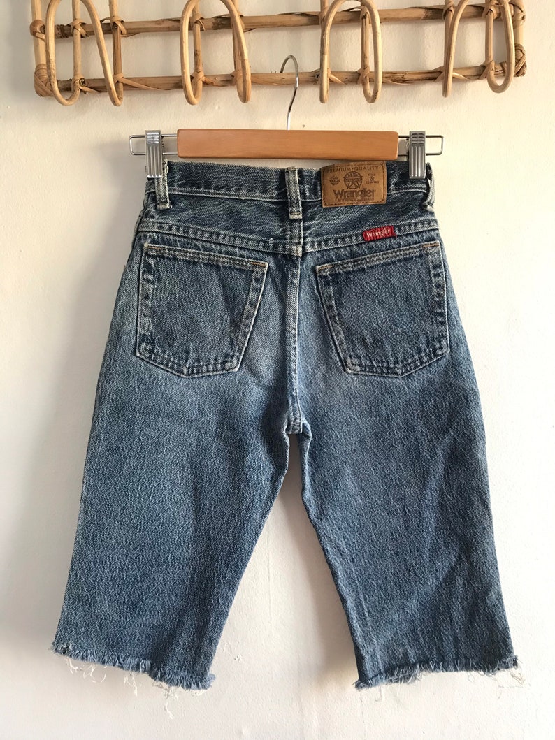 May include: A pair of light blue denim shorts with a frayed hem. The shorts have a brown leather patch on the back with the text "Premium Quality Wrangler" in white.