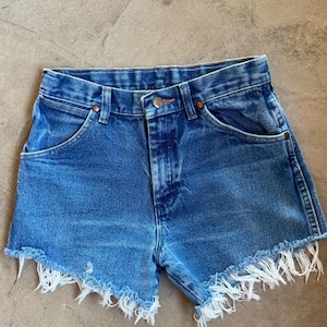 May include: Blue denim shorts featuring a high-waisted design with a button and zipper fly closure, belt loops, and two front pockets. The leg openings are finished with a raw, frayed hemline that exposes white threads along the bottom edges.