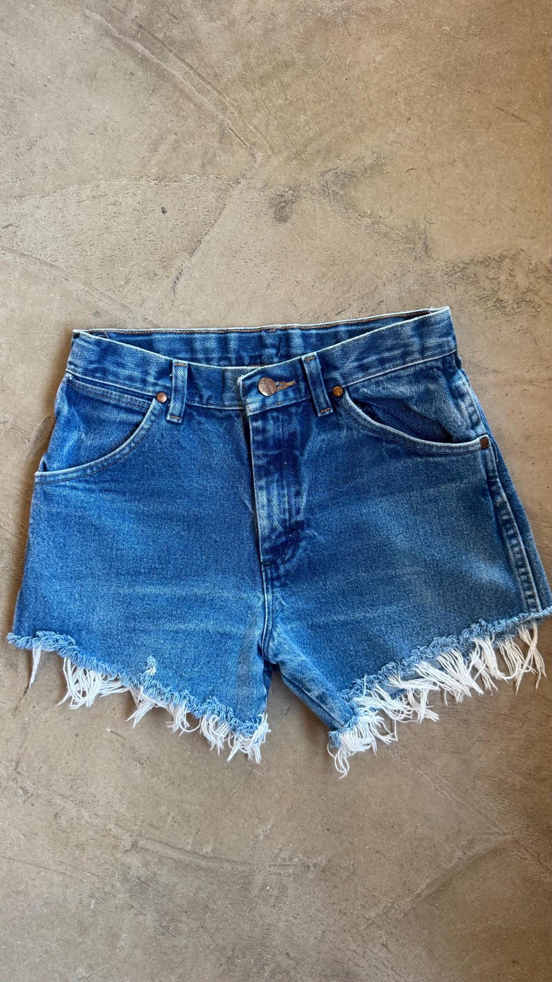 May include: Blue denim shorts featuring a high-waisted design with a button and zipper fly closure, belt loops, and two front pockets. The leg openings are finished with a raw, frayed hemline that exposes white threads along the bottom edges.