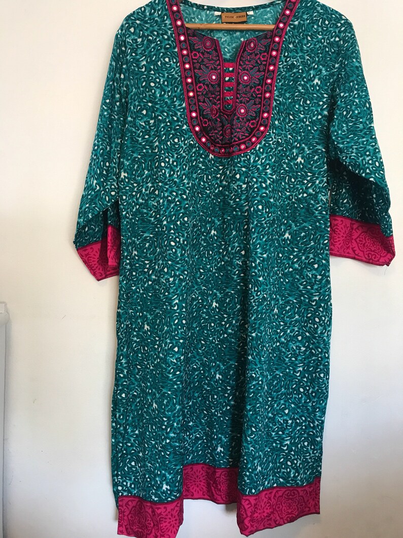Puede incluir: Una t&uacute;nica azul turquesa con estampado floral rosa y blanco. El escote presenta un dise&ntilde;o bordado rosa y plateado con detalles de espejo. La t&uacute;nica tiene mangas tres cuartos y un ribete rosa alrededor del escote y el dobladillo.