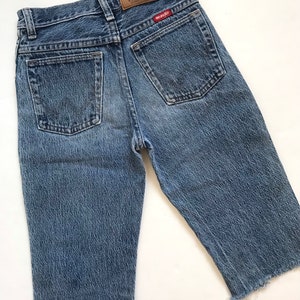 May include: A pair of light blue denim shorts with frayed hems. The shorts have a brown leather patch on the back with the text "Premium Quality Wrangler" and a red Wrangler logo.