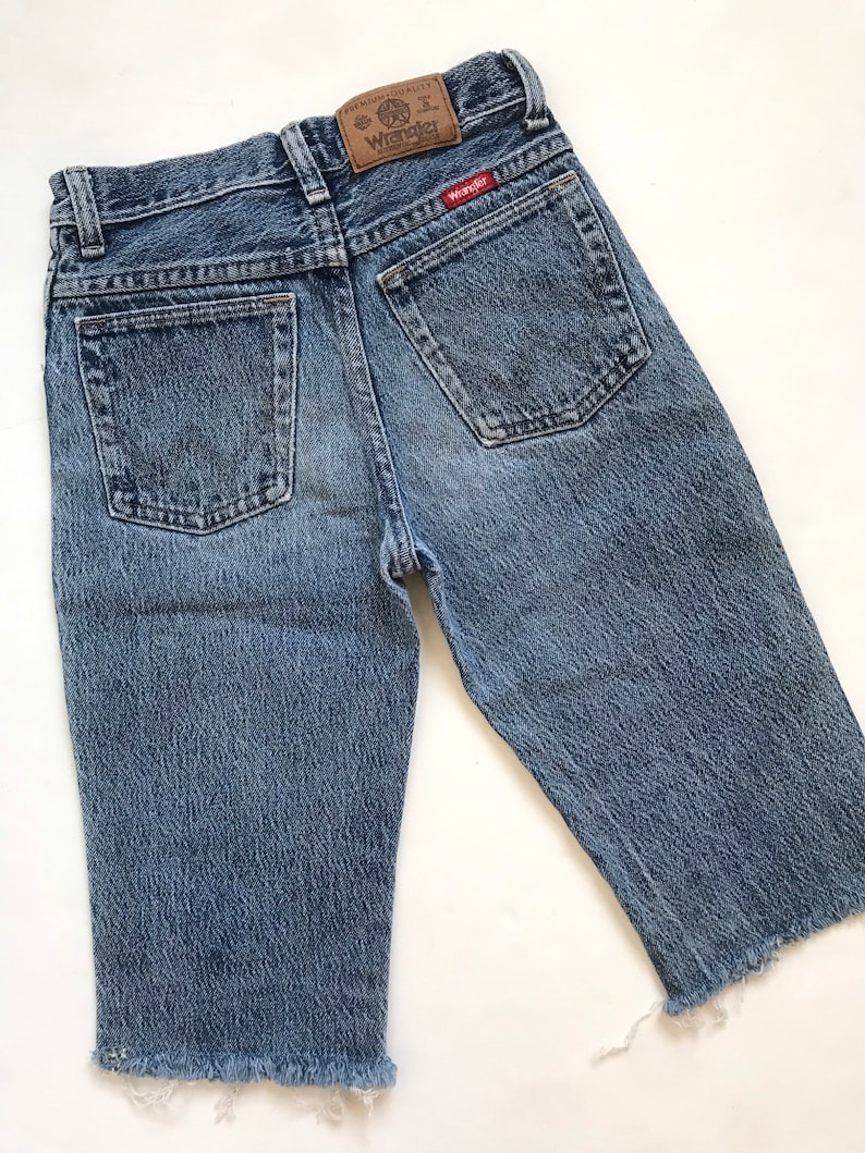 May include: A pair of light blue denim shorts with frayed hems. The shorts have a brown leather patch on the back with the text "Premium Quality Wrangler" and a red Wrangler logo.
