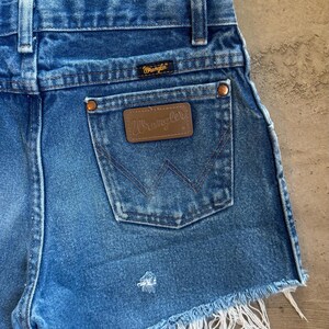May include: Blue denim shorts featuring a high-waisted cut with belt loops, a back pocket with a brown leather Wrangler patch, copper-toned rivets, and a frayed raw hem. The Wrangler logo is also embroidered on a small black tag above the pocket.