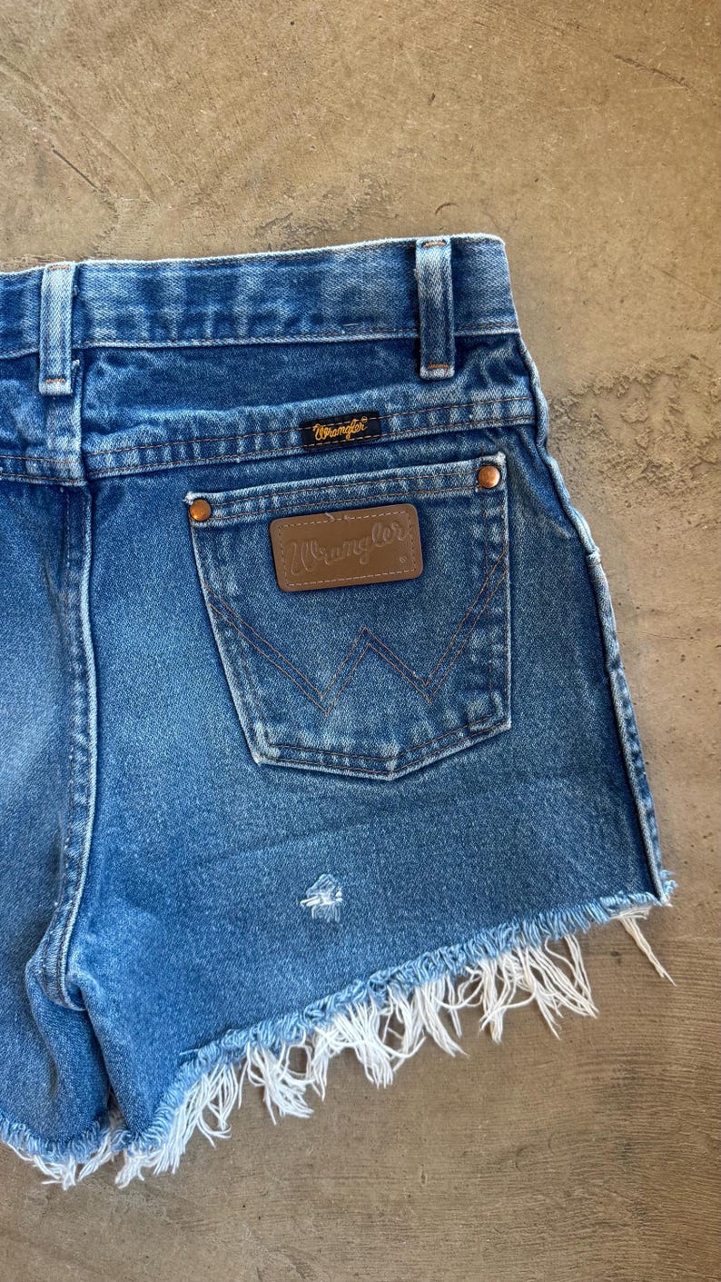 May include: Blue denim shorts featuring a high-waisted cut with belt loops, a back pocket with a brown leather Wrangler patch, copper-toned rivets, and a frayed raw hem. The Wrangler logo is also embroidered on a small black tag above the pocket.