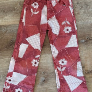 May include: Red patchwork denim pants with white floral designs and a gingham pattern. The pants have a wide leg style.