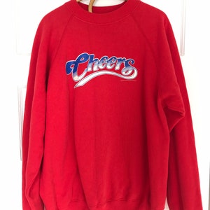 May include: Red sweatshirt with a blue and white "Cheers" graphic on the front.