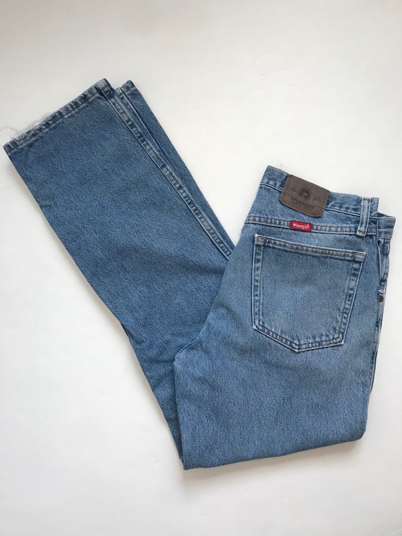 Pu&ograve; includere: Jeans in denim blu chiaro con una toppa in pelle marrone che dice "Wrangler" sulla tasca posteriore.
