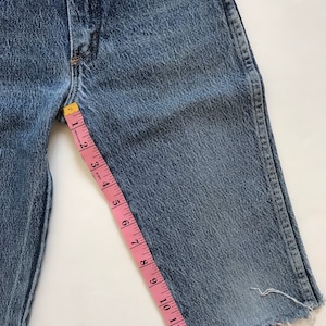 May include: A pair of blue denim shorts with a frayed hem. A pink measuring tape is placed along the side of the shorts, measuring 13 inches.