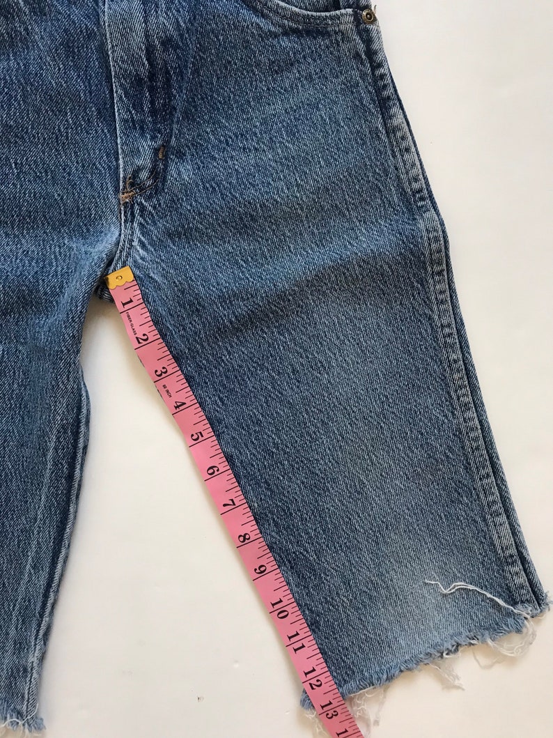 May include: A pair of blue denim shorts with a frayed hem. A pink measuring tape is placed along the side of the shorts, measuring 13 inches.