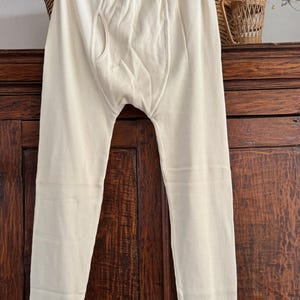 May include: Off-white thermal long underwear pants featuring a ribbed texture, an elastic waistband, and a functional fly opening. The garment has fitted cuffs at the ankles and is constructed from a soft, lightweight knit fabric.