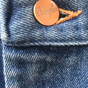 May include: Close-up of a blue denim jean pocket with a copper-colored Wrangler button.