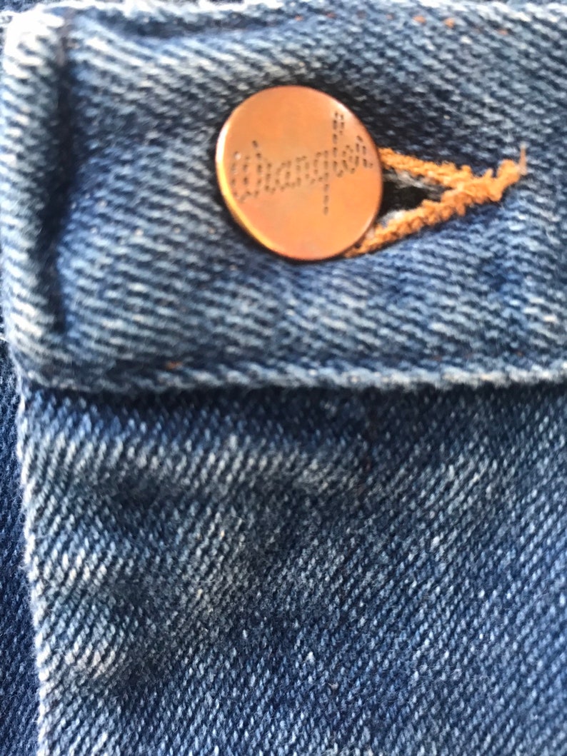 May include: Close-up of a blue denim jean pocket with a copper-colored Wrangler button.