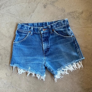 May include: High-waisted blue denim shorts featuring a classic five-pocket design with copper-toned rivets and a button fly closure. The hem is finished with a raw, frayed edge that displays loose white threads along the bottom of the legs.