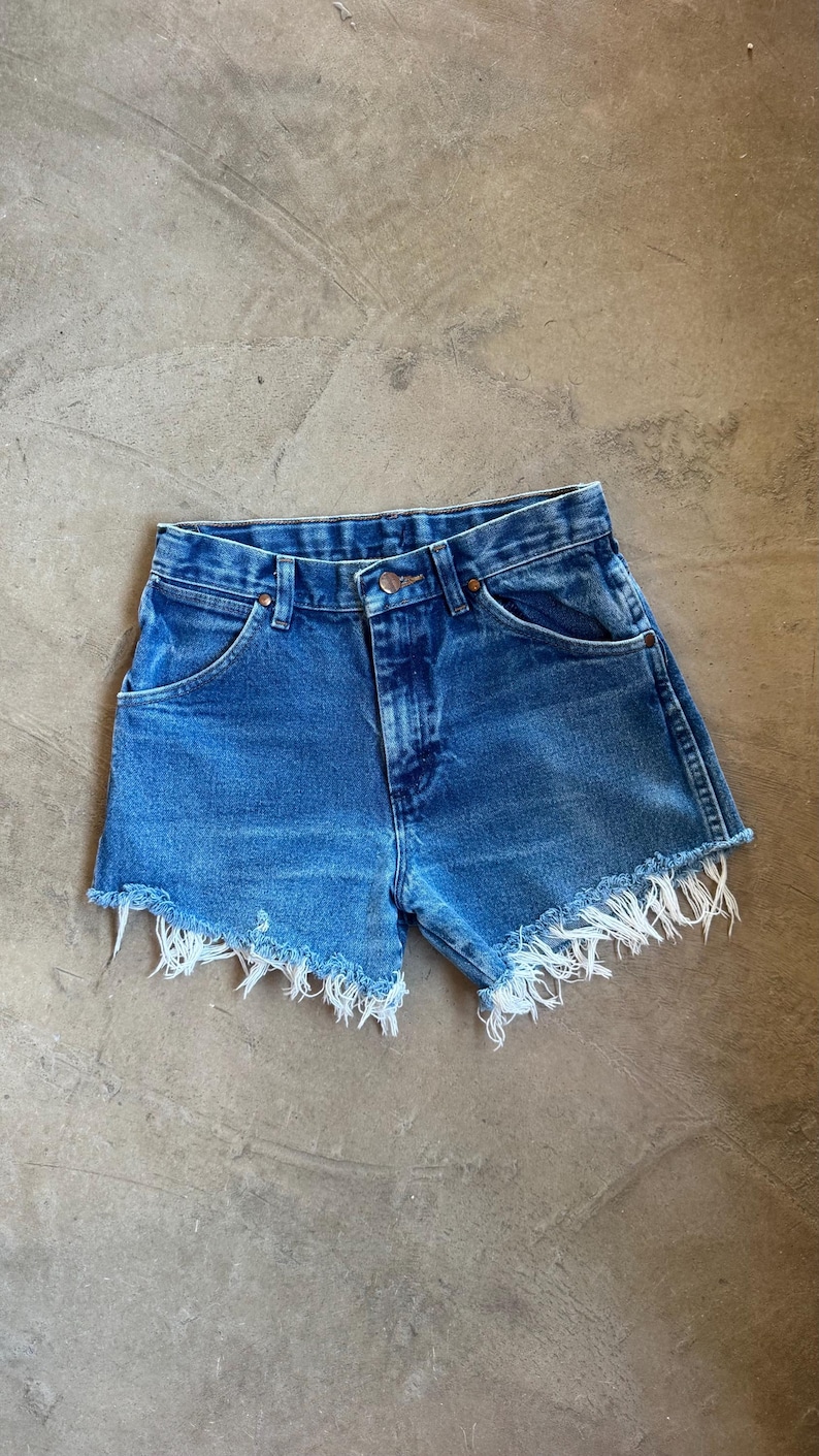 May include: High-waisted blue denim shorts featuring a classic five-pocket design with copper-toned rivets and a button fly closure. The hem is finished with a raw, frayed edge that displays loose white threads along the bottom of the legs.