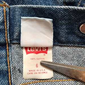K&ouml;nnte beinhalten: Nahaufnahme eines Levi's-Jeans-Etiketts. Das Etikett ist wei&szlig; mit blauer und roter Schrift. Es steht "Levi's" in Rot, "100% Baumwolle" in Blau und "6" in Blau. Das Etikett tr&auml;gt au&szlig;erdem den Hinweis "Hergestellt in den USA. Pflegehinweise auf der R&uuml;ckseite."