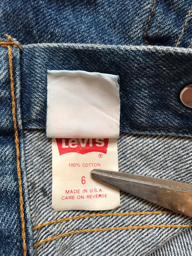 K&ouml;nnte beinhalten: Nahaufnahme eines Levi's-Jeans-Etiketts. Das Etikett ist wei&szlig; mit blauer und roter Schrift. Es steht "Levi's" in Rot, "100% Baumwolle" in Blau und "6" in Blau. Das Etikett tr&auml;gt au&szlig;erdem den Hinweis "Hergestellt in den USA. Pflegehinweise auf der R&uuml;ckseite."