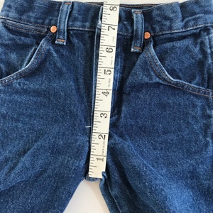 May include: Close-up of blue denim jeans with a measuring tape showing a 2-inch inseam. The jeans have classic five-pocket styling with copper rivets and a button closure. The denim is a medium wash.