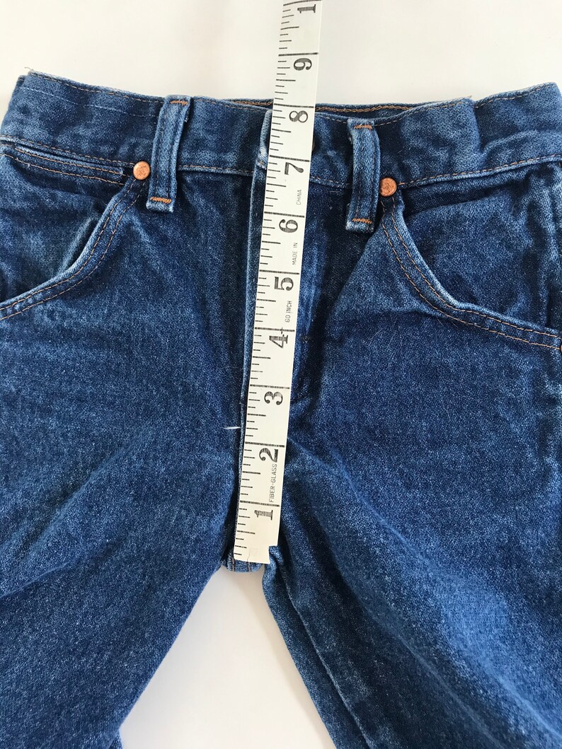 May include: Close-up of blue denim jeans with a measuring tape showing a 2-inch inseam. The jeans have classic five-pocket styling with copper rivets and a button closure. The denim is a medium wash.