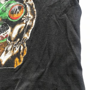 May include: Black t-shirt with a graphic print of a skull wearing green goggles. The skull has a white and orange design.