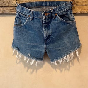 May include: Blue denim shorts featuring a classic five-pocket design, a button fly closure with copper-toned rivets, and a raw, frayed hemline with visible white threads. The medium-wash fabric shows subtle fading and whiskering details.