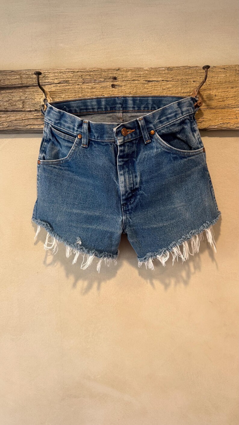 May include: Blue denim shorts featuring a classic five-pocket design, a button fly closure with copper-toned rivets, and a raw, frayed hemline with visible white threads. The medium-wash fabric shows subtle fading and whiskering details.