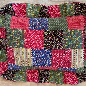 May include: A rectangular quilted pillow featuring a patchwork design of various floral and geometric patterns in shades of red, green, blue, and black. The pillow is finished with a ruffled border made from the same patchwork fabric.